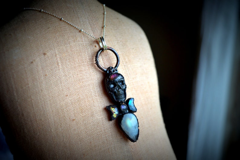 May include: A silver chain necklace with a black skull pendant accented with iridescent moonstone and a pink gemstone. The pendant is attached to a silver ring.