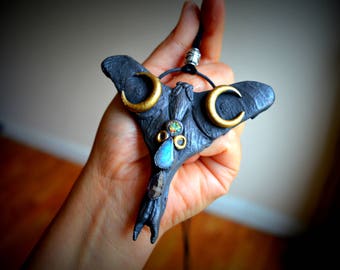 May include: A black bat-shaped pendant with gold crescent moons and a blue gemstone. The pendant is hanging from a black cord.