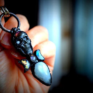 May include: A black skull pendant with a silver ring and a variety of colorful gemstones, including a large iridescent moonstone.
