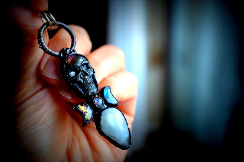 May include: A black skull pendant with a silver ring and a variety of colorful gemstones, including a large iridescent moonstone.
