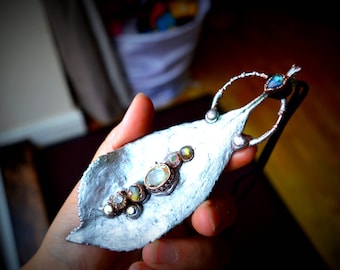 May include: A silver leaf-shaped pendant with a copper wire frame and five iridescent gemstones. The pendant is held in a hand.