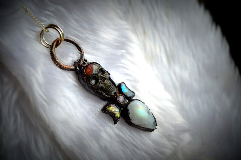 May include: A silver chain necklace with a copper pendant featuring a skull, moonstone, and iridescent stones.