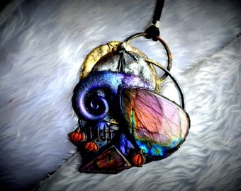 May include: A handcrafted pendant necklace featuring a skull design with a large, iridescent stone. The pendant has a dark blue, purple, and gold color scheme, with small orange pumpkin accents. The necklace hangs on a black cord.
