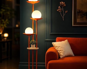 May include: A tall orange floor lamp featuring three vertical poles of varying heights, each topped with a white spherical glass shade. A small circular shelf holds a potted green plant, and the base is a solid orange disc.