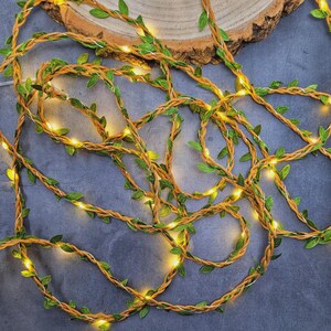 May include: A string of artificial ivy vine lights featuring a flexible brown cord intertwined with small green fabric leaves and warm white LED bulbs. The decorative garland is arranged in a loose, coiled pattern, showcasing the illuminated foliage.