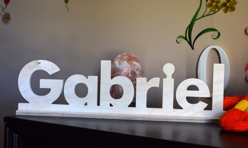 Puede incluir: Letras de madera blancas que deletrean el nombre "Gabriel" sobre una base de madera. Las letras est&aacute;n recortadas y se encuentran sobre un estante.