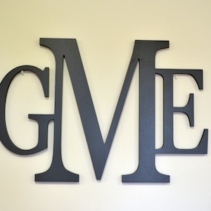 May include: Black wooden letters spelling out the word "GME" mounted on a light beige wall.