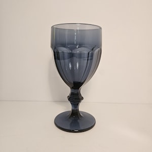 May include: A smoke-gray glass goblet featuring a smooth, wide rim and a bowl decorated with vertical, rounded fluted panels. The stem is faceted with a central decorative node, leading down to a circular, flat base for stability.