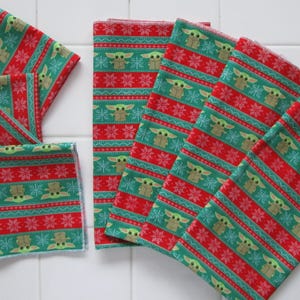 Puede incluir: Una colecci&oacute;n de servilletas de tela estampadas en rojo y verde. El dise&ntilde;o presenta un patr&oacute;n repetitivo de un personaje, copos de nieve y rayas. Las servilletas est&aacute;n dispuestas sobre una superficie de baldosas blancas.