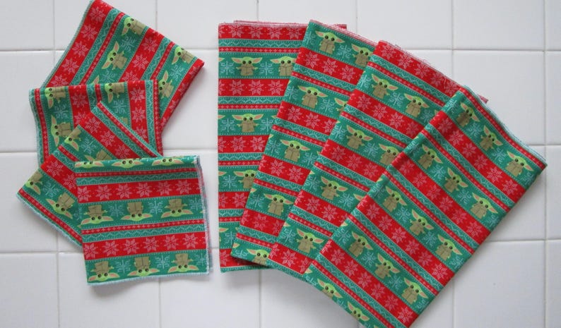 Puede incluir: Una colecci&oacute;n de servilletas de tela estampadas en rojo y verde. El dise&ntilde;o presenta un patr&oacute;n repetitivo de un personaje, copos de nieve y rayas. Las servilletas est&aacute;n dispuestas sobre una superficie de baldosas blancas.