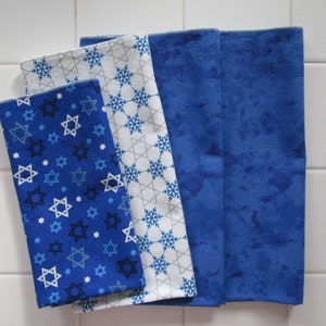 May include: Four blue and white fabric napkins with different patterns. One napkin has a blue background with white stars of David, another has a white background with blue snowflakes, and the other two have a blue background with a white marbled pattern.