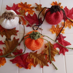 May include: Three crocheted pumpkin-shaped drawstring bags in white, orange, and red, surrounded by artificial fall leaves.