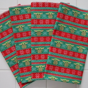 Puede incluir: Cuatro servilletas de tela estampadas en rojo y verde. Las servilletas presentan un patr&oacute;n repetido de un personaje de dibujos animados y copos de nieve. El fondo es rojo con rayas verdes. Las servilletas est&aacute;n dobladas y apiladas.