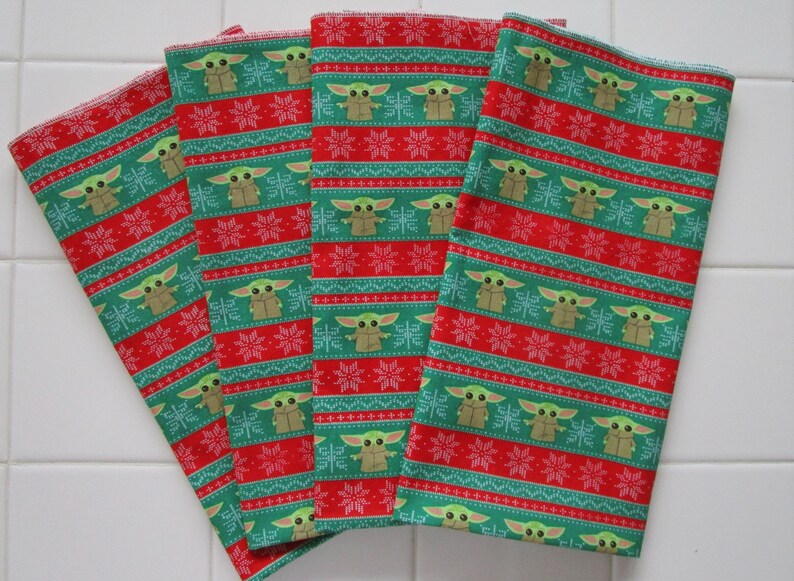 Puede incluir: Cuatro servilletas de tela estampadas en rojo y verde. Las servilletas presentan un patr&oacute;n repetido de un personaje de dibujos animados y copos de nieve. El fondo es rojo con rayas verdes. Las servilletas est&aacute;n dobladas y apiladas.