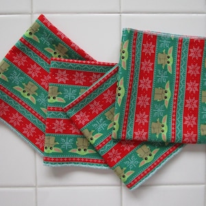 Puede incluir: Cuatro servilletas de tela cuadradas con un patr&oacute;n festivo navide&ntilde;o. Las servilletas presentan un dise&ntilde;o repetitivo de un personaje verde, copos de nieve y rayas rojas. Las servilletas son de tela y est&aacute;n dobladas y apiladas sobre una superficie de baldosas blancas.