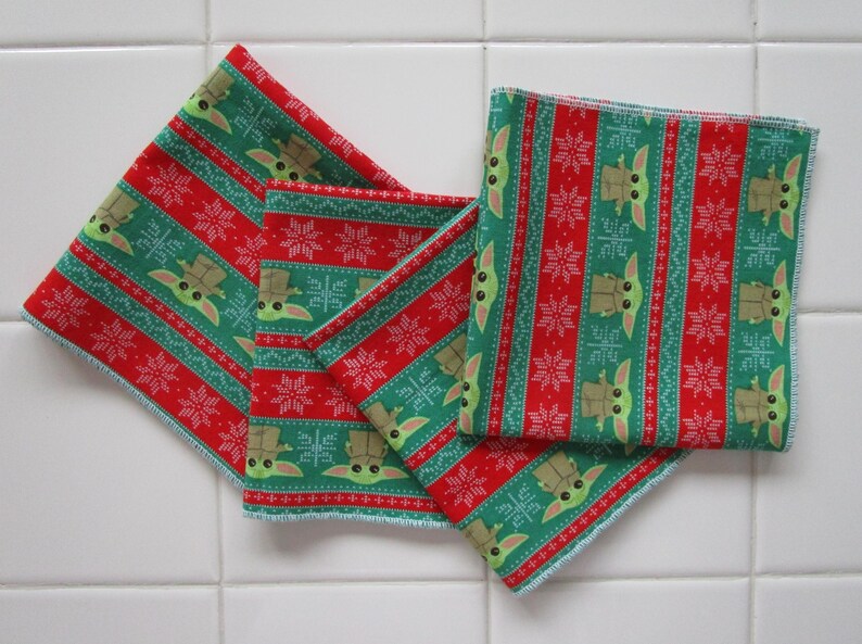 Puede incluir: Cuatro servilletas de tela cuadradas con un patr&oacute;n festivo navide&ntilde;o. Las servilletas presentan un dise&ntilde;o repetitivo de un personaje verde, copos de nieve y rayas rojas. Las servilletas son de tela y est&aacute;n dobladas y apiladas sobre una superficie de baldosas blancas.