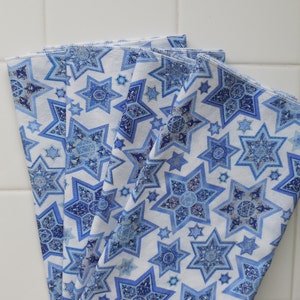 May include: Four white fabric napkins with a blue star of David pattern. The stars are detailed with a glittery, iridescent finish.