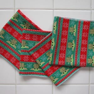 Puede incluir: Cuatro cuadrados de tela rojos y verdes con un patr&oacute;n repetido de un personaje y copos de nieve. Los cuadrados est&aacute;n dispuestos en forma de abanico, con los bordes de la tela visibles. La tela tiene un tema festivo y navide&ntilde;o.