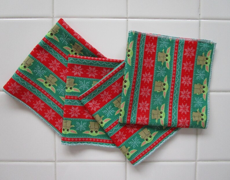 Puede incluir: Cuatro cuadrados de tela rojos y verdes con un patr&oacute;n repetido de un personaje y copos de nieve. Los cuadrados est&aacute;n dispuestos en forma de abanico, con los bordes de la tela visibles. La tela tiene un tema festivo y navide&ntilde;o.
