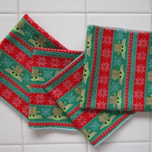 Puede incluir: Cuatro cuadrados de tela estampada en verde y rojo. El patr&oacute;n presenta un dise&ntilde;o repetitivo de un personaje con ojos y orejas grandes, junto con copos de nieve. Los bordes de los cuadrados est&aacute;n acabados con costuras azul claro.