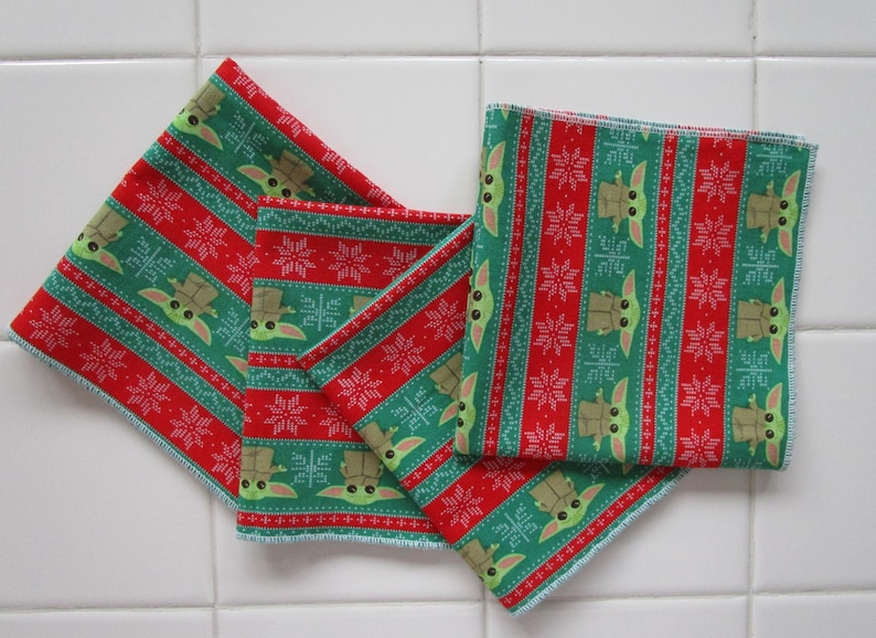 Puede incluir: Cuatro cuadrados de tela estampada en verde y rojo. El patr&oacute;n presenta un dise&ntilde;o repetitivo de un personaje con ojos y orejas grandes, junto con copos de nieve. Los bordes de los cuadrados est&aacute;n acabados con costuras azul claro.