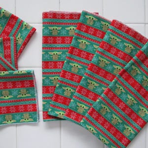 Puede incluir: Una colecci&oacute;n de cuadrados de tela estampada en rojo y verde. La tela presenta un dise&ntilde;o repetitivo de un personaje, copos de nieve y rayas. Los cuadrados est&aacute;n dispuestos en forma de abanico, algunos doblados y apilados. El tema general es festivo y navide&ntilde;o.