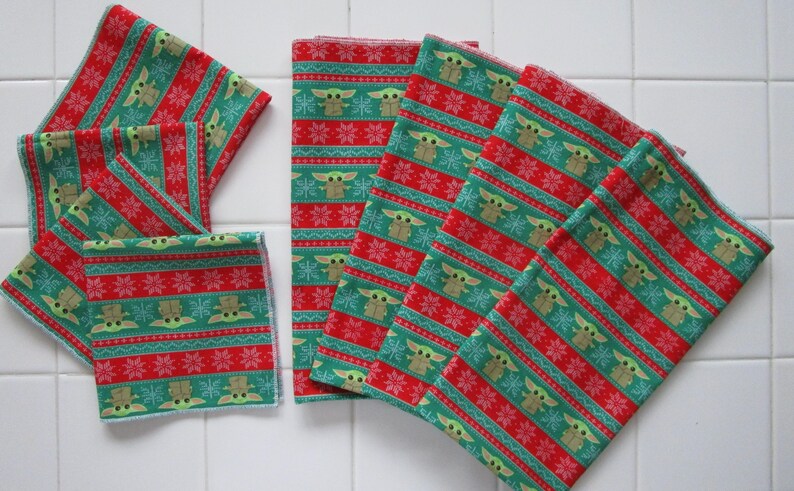 Puede incluir: Una colecci&oacute;n de cuadrados de tela estampada en rojo y verde. La tela presenta un dise&ntilde;o repetitivo de un personaje, copos de nieve y rayas. Los cuadrados est&aacute;n dispuestos en forma de abanico, algunos doblados y apilados. El tema general es festivo y navide&ntilde;o.