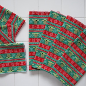 Puede incluir: Una colecci&oacute;n de cuadrados de tela estampada en verde, rojo y blanco. El patr&oacute;n presenta un dise&ntilde;o repetitivo de un personaje con ojos y orejas grandes, copos de nieve y rayas. Los cuadrados de tela est&aacute;n dispuestos sobre una superficie de baldosas blancas.