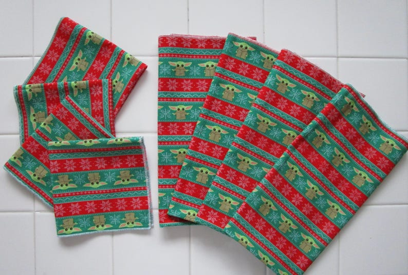 Puede incluir: Una colecci&oacute;n de cuadrados de tela estampada en verde, rojo y blanco. El patr&oacute;n presenta un dise&ntilde;o repetitivo de un personaje con ojos y orejas grandes, copos de nieve y rayas. Los cuadrados de tela est&aacute;n dispuestos sobre una superficie de baldosas blancas.