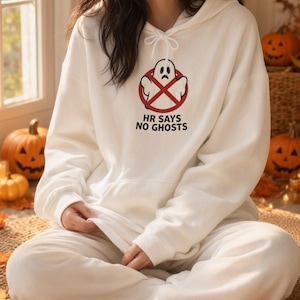 Peut inclure: Un sweat-shirt à capuche blanc orné d'un motif représentant un fantôme dans un cercle rouge barré d'une ligne diagonale. Sous le motif, le texte HR SAYS NO GHOSTS est inscrit en lettres capitales noires. Le sweat possède des manches longues.