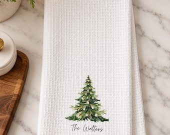 May include: A white waffle-weave kitchen towel featuring a watercolor illustration of a decorated Christmas tree with lights and ornaments, with the text The Walters printed in black script lettering below the tree design.