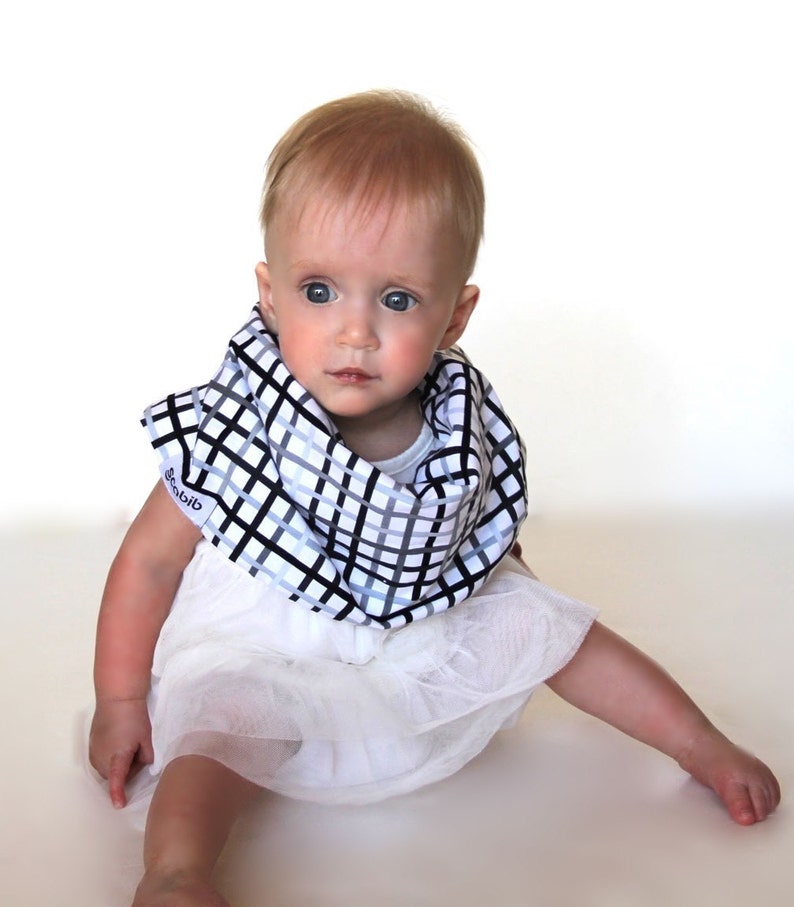May include: A baby wearing a black, white, and gray checkered bib. The bib is around the baby's neck, and the baby is wearing a white dress. The bib has the word "Scobib" printed on it.