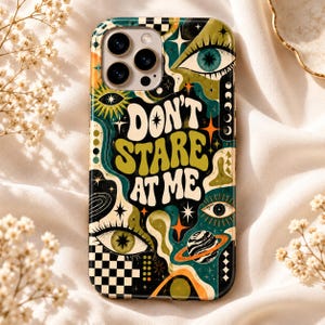 May include: A phone case featuring a dark green, olive, and orange graphic design with the text DON'T STARE AT ME. The pattern includes multiple eyes, stars, a planet with rings, and a black and white checkerboard section at the bottom.