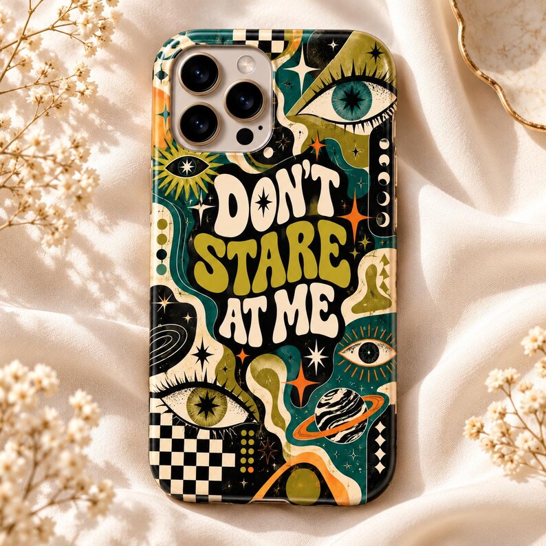 May include: A phone case featuring a dark green, olive, and orange graphic design with the text DON'T STARE AT ME. The pattern includes multiple eyes, stars, a planet with rings, and a black and white checkerboard section at the bottom.
