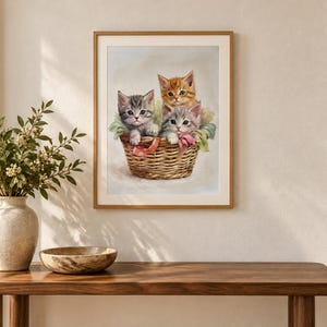 May include: A framed wall art print featuring three kittens sitting inside a woven wicker basket. The kittens include one gray tabby, one orange tabby, and one light gray kitten, all nestled together with soft fabric accents inside the basket.