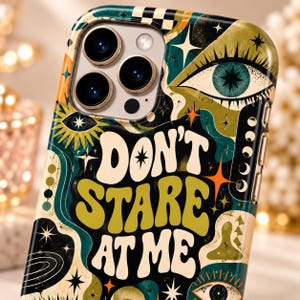 May include: A smartphone case featuring a dark green, cream, and orange graphic pattern with multiple eye illustrations, star shapes, and the text "DON'T STARE AT ME" in a bold, wavy font. The case has a cutout for a triple-lens camera system.