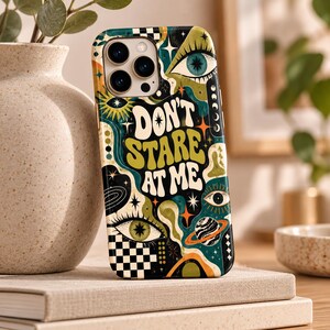 May include: A smartphone case featuring a psychedelic design with the text DON'T STARE AT ME in bold lettering. The graphic includes multiple eyes, a planet with rings, stars, and a black and white checkerboard pattern in shades of green, orange, and black.