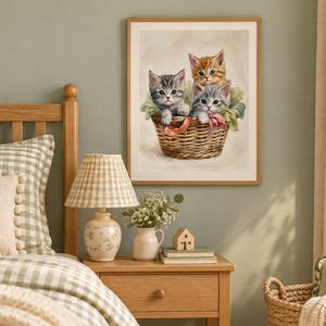 May include: A framed wall art print featuring a painting of three kittens, one orange tabby and two gray tabby kittens, sitting inside a woven wicker basket. The kittens are nestled among soft green fabric and pink ribbons.