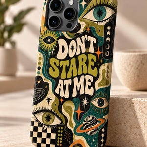 May include: A smartphone case featuring a dark background with green, orange, and cream-colored graphic elements including multiple eyes, stars, a planet with rings, and a checkerboard pattern. The text DON'T STARE AT ME is printed in large, bold, wavy cream letters.