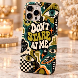 May include: A smartphone case featuring a dark background with green, orange, and white graphic patterns including eyes, stars, a planet with rings, and a checkerboard design. The text DON'T STARE AT ME is printed in large, bold, wavy white letters.
