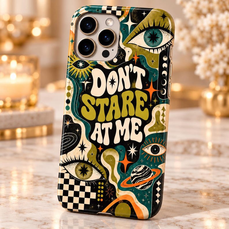 May include: A smartphone case featuring a dark background with green, orange, and white graphic patterns including eyes, stars, a planet with rings, and a checkerboard design. The text DON'T STARE AT ME is printed in large, bold, wavy white letters.