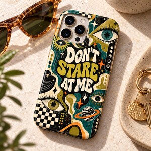 May include: A smartphone case featuring a black, green, and orange graphic design with the text "DON'T STARE AT ME". The pattern includes stylized eyes, stars, a planet with rings, and a checkered border, all set against a dark background.