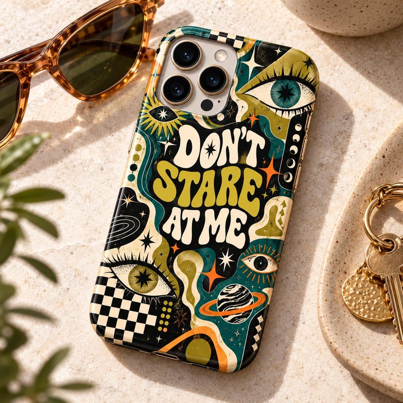May include: A smartphone case featuring a black, green, and orange graphic design with the text "DON'T STARE AT ME". The pattern includes stylized eyes, stars, a planet with rings, and a checkered border, all set against a dark background.