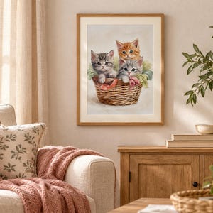 May include: A framed wall art print featuring a painting of three kittens sitting inside a woven wicker basket. One kitten is orange tabby, while the other two are gray tabby, all nestled together with soft green fabric accents inside the basket.