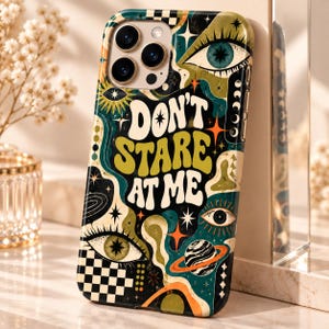 May include: A smartphone case featuring a dark teal, olive green, and orange graphic design with the text "DON'T STARE AT ME" in a bold, wavy font. The pattern includes multiple stylized eyes, stars, a planet with rings, and a black and white checkerboard.