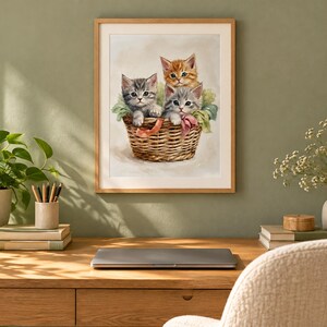 May include: A framed wall art print featuring a watercolor illustration of three kittens sitting inside a woven wicker basket. One kitten is orange tabby, while the other two are gray tabby, all nestled among soft green and pink fabric accents.