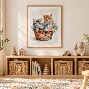 May include: A framed wall art print featuring a painting of three kittens, one orange tabby and two gray tabbies, sitting inside a woven wicker basket with colorful ribbons. The artwork is displayed in a light wood frame on a neutral wall.