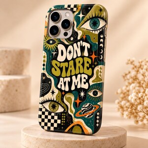 May include: A smartphone case featuring a dark background with a colorful graphic print of eyes, stars, a planet with rings, and a checkerboard pattern. The text "DON'T STARE AT ME" is printed in large, bold, cream-colored letters in the center.