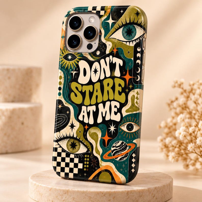 May include: A smartphone case featuring a dark background with a colorful graphic print of eyes, stars, a planet with rings, and a checkerboard pattern. The text "DON'T STARE AT ME" is printed in large, bold, cream-colored letters in the center.