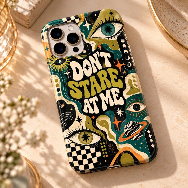 May include: A smartphone case featuring a dark green, black, and cream graphic design with the text "DON'T STARE AT ME" in a bold, wavy font. The pattern includes multiple eyes, stars, a planet with rings, and a black and white checkerboard.